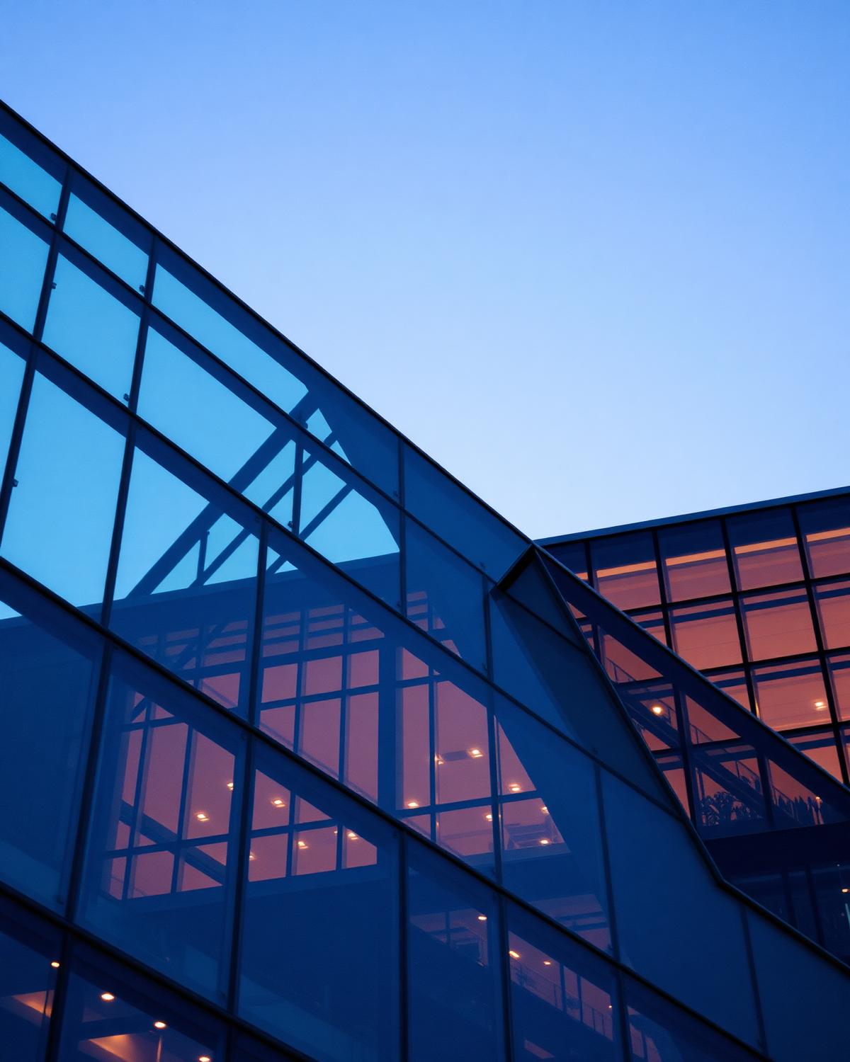Angled glass and steel facade of a modern building at dusk with warm interior lighting